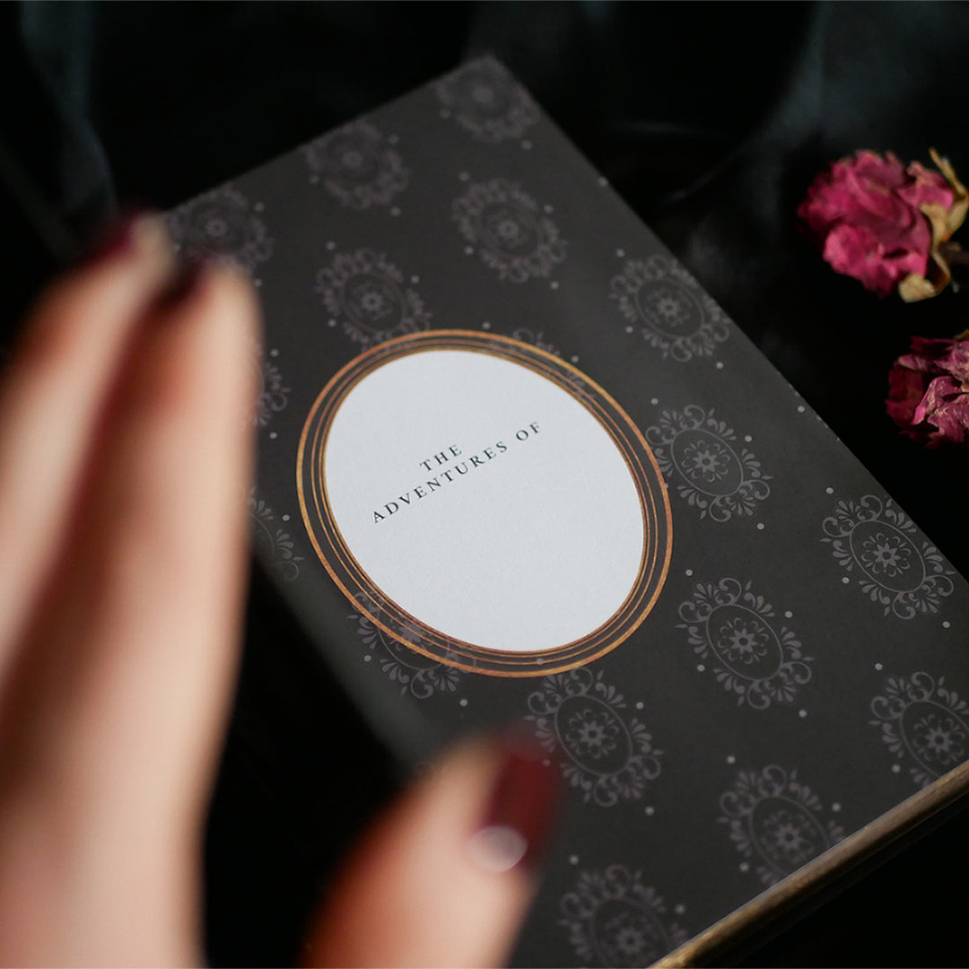 A close-up of a Black Literati Journal by The Quirky Cup Collective, featuring a patterned cover with an ornate design and a central white oval that reads 'The Adventures Of.' A hand with dark red nail polish is slightly blurred in the foreground, suggesting movement or anticipation. The journal rests on a dark fabric background, with dried pink roses placed to the side, adding a touch of vintage elegance to the composition.