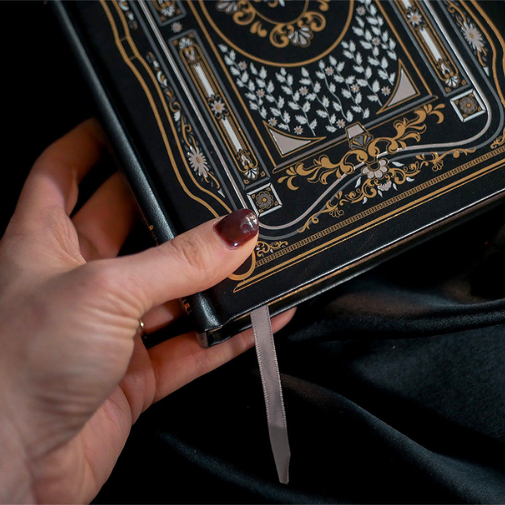 A close-up of a Black Literati Journal by The Quirky Cup Collective, featuring a patterned cover with an ornate design. A hand with dark red nail polish is slightly blurred in the foreground, suggesting movement or anticipation. The journal rests on a dark fabric background, with dried pink roses placed to the side, adding a touch of vintage elegance to the composition.
