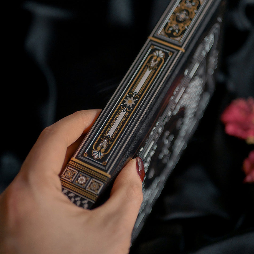 A close-up of a Black Literati Journal by The Quirky Cup Collective, featuring a patterned cover with an ornate design. A hand with dark red nail polish is slightly blurred in the foreground, suggesting movement or anticipation. The journal rests on a dark fabric background, with dried pink roses placed to the side, adding a touch of vintage elegance to the composition.
