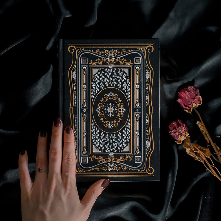 A hand holding the Black Literati Journal by The Quirky Cup Collective, showcasing its intricate gold and white floral and geometric cover design on a black background. The journal is positioned on top of a dark, silky fabric, with dried pink roses placed next to it, creating a moody, vintage-inspired scene. The journal's ribbon bookmark is partially visible, adding a touch of elegance to the overall aesthetic.
