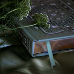 The sage green literati Journal sits on a sage green background surrounded by white and green flowers. The journal features gold gilded page edges and an  intricate gold, green and white foiling with it's cover design inspired by classical books and a love of reading.