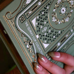 a close up of  back of the sage green Journal showing the logo. The journal features gold gilded page edges and an  intricate gold, green and white foiling with it's cover design inspired by classical books and a love of reading.