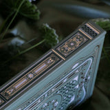 The spine of the sage green Literati Journal shows the intricate gold, green and white design that is foiled. The geometric floral design is inspired by classical books and a love of reading. The book sits on a sage green fabric background surrounded by flowers.