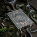 The sage green Literati Journal sits on a green fabric background surrounded by flowers. The journal features intricate gold, green and white foiling of a design inspired by classical books and a love of reading. The journal also features lush gold gilded pages.