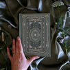 The sage green Literati Journal is held by a hand on a green fabric background surrounded by flowers. The journal features intricate gold, green and white foiling of a design inspired by classical books and a love of reading.