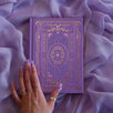 The soft lilac journal as part of the classic book inspired Wisteria Literati collection sits on a light purple fabric background. This soft purple journal features a slightly Victorian book inspired motif in shades of gold, white, pink and purple alongside intricate foil detailing.
