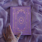 The soft lilac journal as part of the classic book inspired Wisteria Literati collection sits on a light purple fabric background. This soft purple journal features a slightly Victorian book inspired motif in shades of gold, white, pink and purple alongside intricate foil detailing.