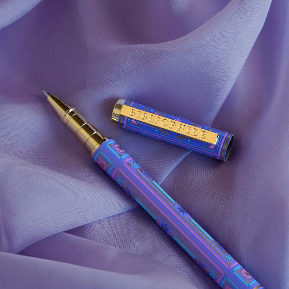 Detail image of the wisteria lilac Bibliophile pen by The Quirky Cup Collective. The light purple pen features a classic book inspired design in shades of pink, purple and blue with gold detailing. The gold pen clip features an engraving of the phrase 'Bibliophile'. The pen sits on a light purple fabric background.