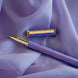 Detail image of the wisteria lilac Bibliophile pen by The Quirky Cup Collective. The light purple pen features a classic book inspired design in shades of pink, purple and blue with gold detailing. The gold pen clip features an engraving of the phrase 'Bibliophile'. The pen sits on a light purple fabric background.