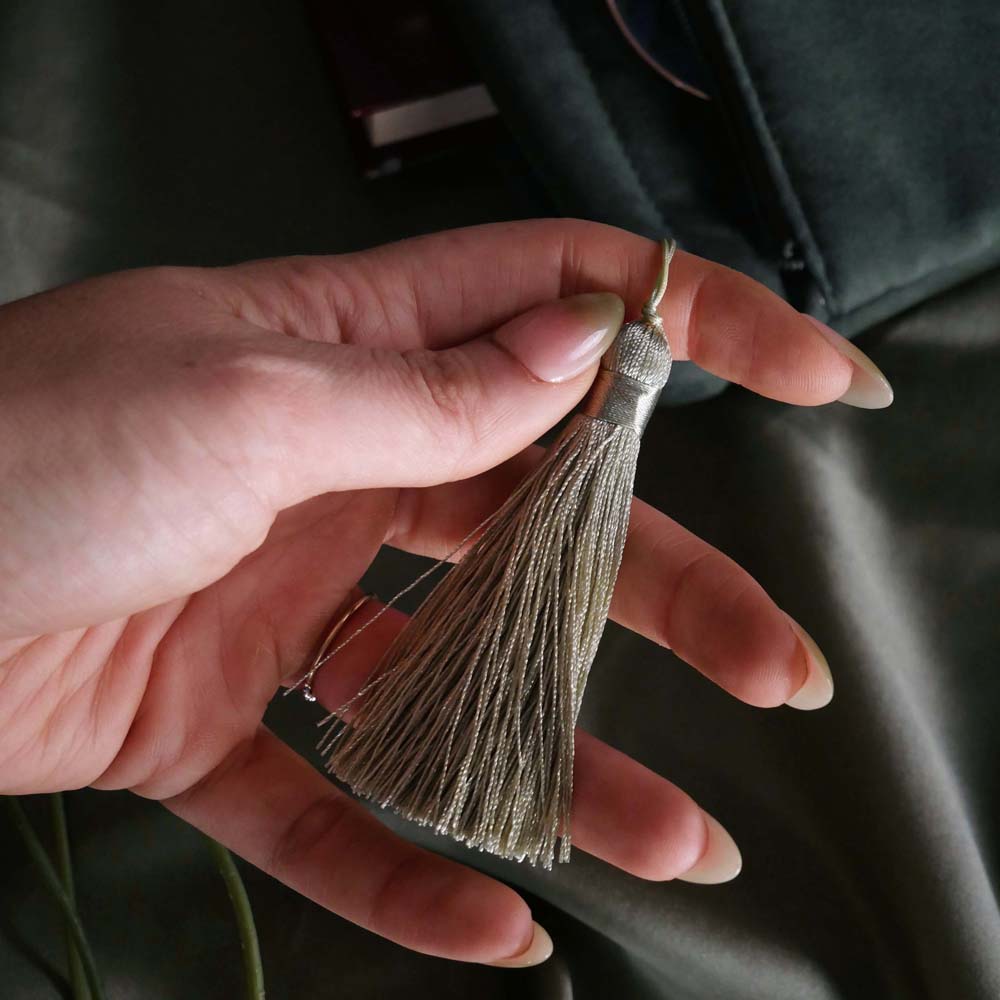 The detail of the sage green literati book sleeve by The Quirky Cup Collective. The soft green velvet case features intricate gold, white and light green embroidery in the design of a classical book motif. The book case features a soft green tassel and sits on a light green fabric background.