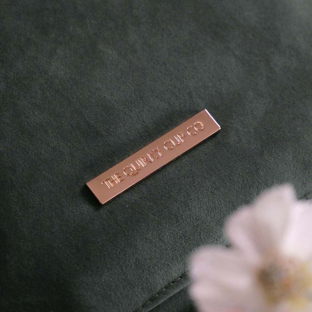 The sage green literati book sleeve by The Quirky Cup Collective. The soft green velvet case features intricate gold, white and light green embroidery in the design of a classical book motif. The book case features a soft green tassel and sits on a light green fabric background. Here the gold logo badge on the back is shown.