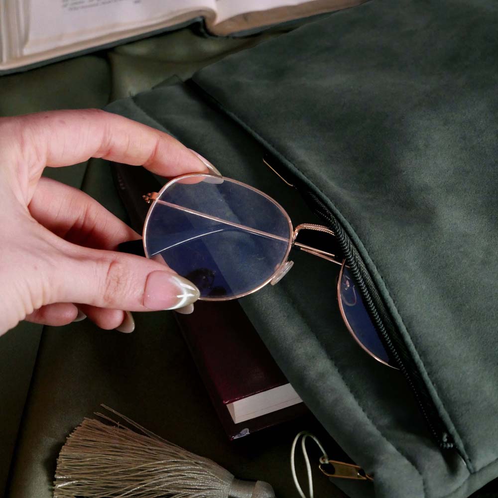 Hand putting a pair of sunglasses into into the sage green literati book sleeve pocket by The Quirky Cup Collective. The soft green velvet case features intricate gold, white and light green embroidery in the design of a classical book motif. The book case features a soft green tassel and sits on a light green fabric background.