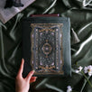 The sage green literati book sleeve by The Quirky Cup Collective. The soft green velvet case features intricate gold, white and light green embroidery in the design of a classical book motif. The book case features a soft green tassel and sits on a light green fabric background, a small Burgundy book poking out the top.