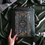 The sage green literati book sleeve by The Quirky Cup Collective. The soft green velvet case features intricate gold, white and light green embroidery in the design of a classical book motif. The book case features a soft green tassel and sits on a light green fabric background, a small Burgundy book poking out the top.