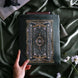 The sage green literati book sleeve by The Quirky Cup Collective. The soft green velvet case features intricate gold, white and light green embroidery in the design of a classical book motif. The book case features a soft green tassel and sits on a light green fabric background, a small Burgundy book poking out the top.