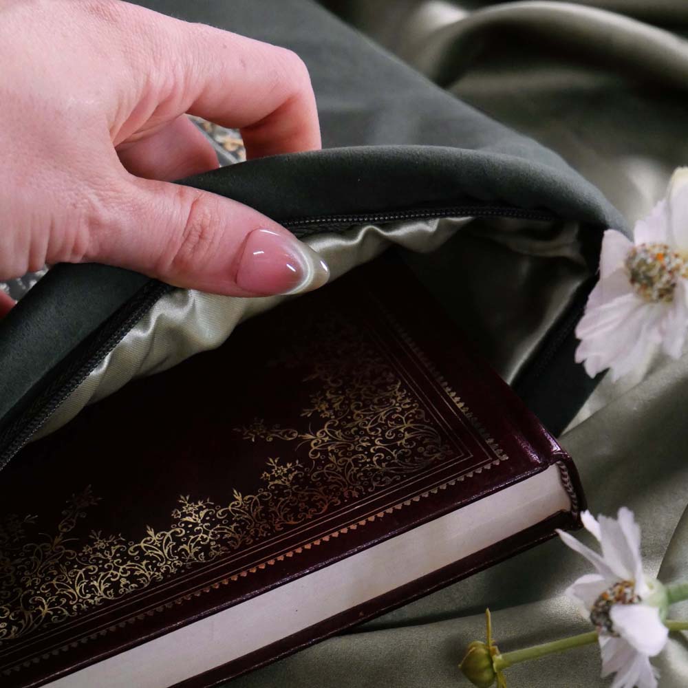 Hand opening the sage green literati book sleeve by The Quirky Cup Collective as it holds an ornate book design. The soft green velvet case features intricate gold, white and light green embroidery in the design of a classical book motif. The book case features a soft green tassel and sits on a light green fabric background.