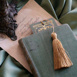 The top edge of the sage green literati bookmark peeks out from a green book. the bookmark features a Victorian book inspired design with intricate gold foiling and a soft gold tassel. 