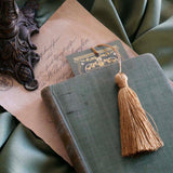 The top edge of the sage green literati bookmark peeks out from a green book. the bookmark features a Victorian book inspired design with intricate gold foiling and a soft gold tassel. 