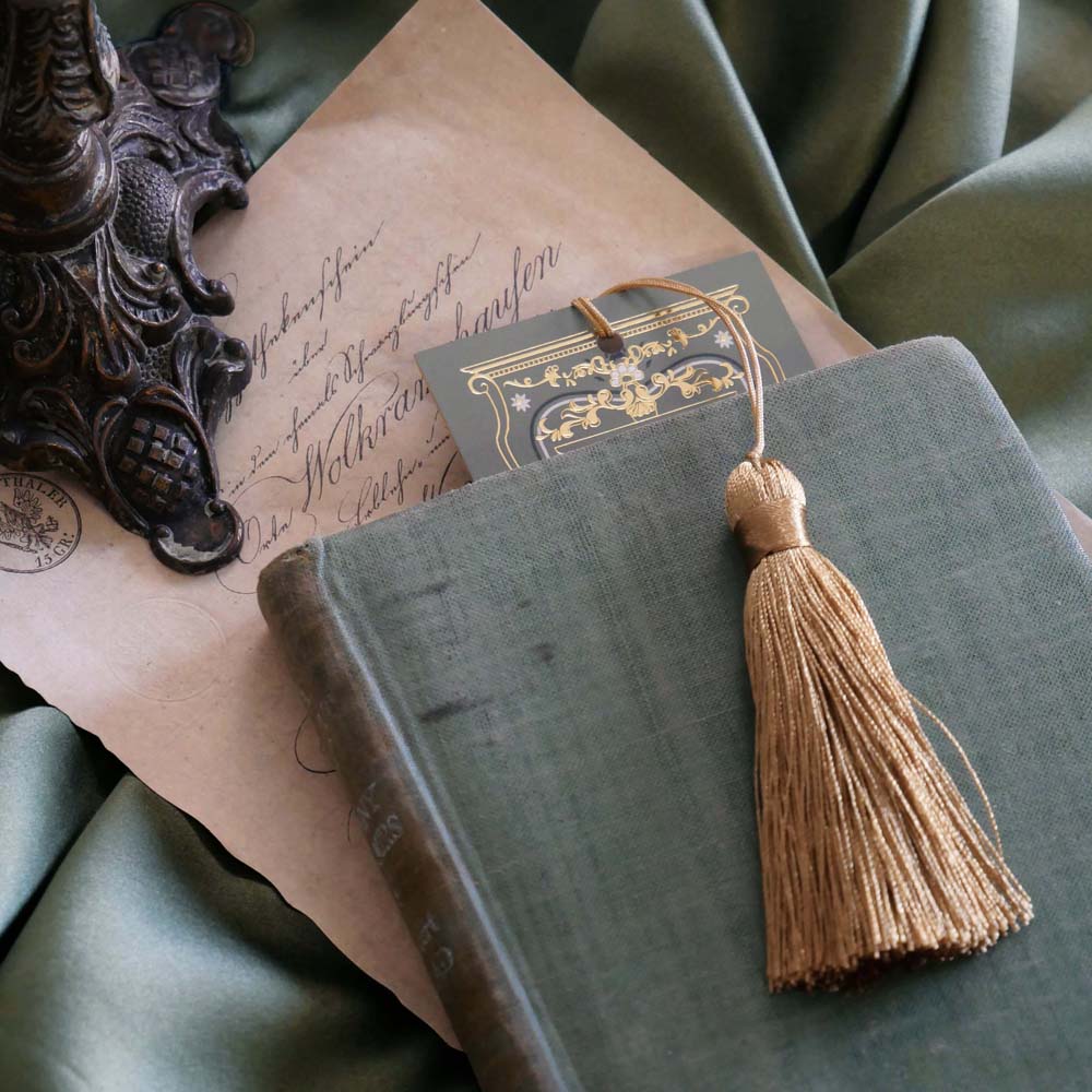 The top edge of the sage green literati bookmark peeks out from a green book. the bookmark features a Victorian book inspired design with intricate gold foiling and a soft gold tassel. 