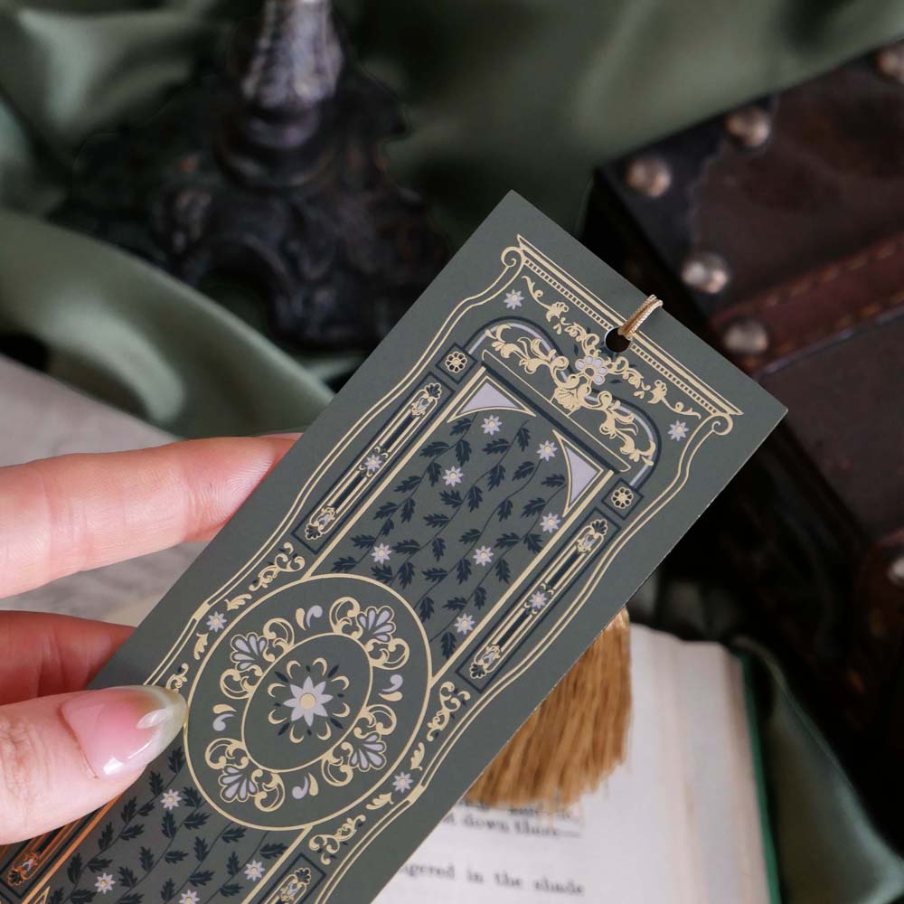 The sage green literati bookmark features a classical book inspired design with intricate gold foiling and a soft gold tassel. The bookmark is held by a hand in front of a soft green fabric background surrounded by classical books.
