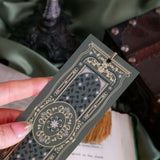 The sage green literati bookmark features a classical book inspired design with intricate gold foiling and a soft gold tassel. The bookmark is held by a hand in front of a soft green fabric background surrounded by classical books.
