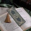 The sage green literati bookmark features a classical book inspired design with intricate gold foiling and a soft gold tassel. The bookmark sits on a classical book on a soft green fabric background.