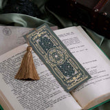 The sage green literati bookmark features a classical book inspired design with intricate gold foiling and a soft gold tassel. The bookmark sits on a classical book on a soft green fabric background.