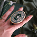 A  hand holds the Sage Literati clingie The kindle grip (similar to an enamel pop socket) features a gold and green floral motif design and is shown in front of a sage green background.