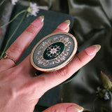 A  hand holds the Sage Literati clingie The kindle grip (similar to an enamel pop socket) features a gold and green floral motif design and is shown in front of a sage green background.