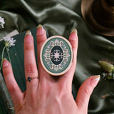 A  hand holds the Sage Literati clingie The kindle grip (similar to an enamel pop socket) features a gold and green floral motif design and is shown in front of a sage green background.