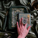 The Sage Literati eReader bundle is shown with a hand holding the gold and green oval shaped eReader grip attatched to a Kindle with matching sage green sticker insert. The kindle grip (similar to an enamel pop socket) is accompanied by a gold Charmlet with dangling charms and a matching sage green eReader sleeve all sitting on a sage green background.