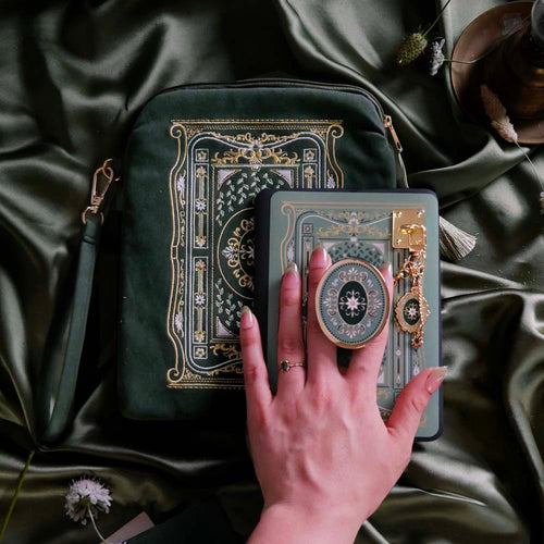 The Sage Literati eReader bundle is shown with a hand holding the gold and green oval shaped eReader grip attatched to a Kindle with matching sage green sticker insert. The kindle grip (similar to an enamel pop socket) is accompanied by a gold Charmlet with dangling charms and a matching sage green eReader sleeve all sitting on a sage green background.