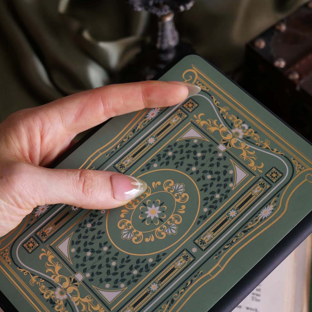 The sage green Literati eReader insert sticker stick to a back kindle. The foiled sticker features a victorian book inspired motif with an intricate design in shades of green, white and gold foil. A hand holds the Kindle in front of a light green fabric background.