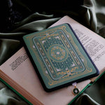 The sage green Literati eReader insert sticker stick to a back kindle. The foiled sticker features a victorian book inspired motif with an intricate design in shades of green, white and gold foil. The Kindle sits on an open classical book on top of a light green fabric background.