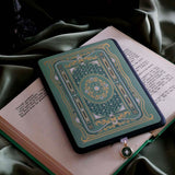 The sage green Literati eReader insert sticker stick to a back kindle. The foiled sticker features a victorian book inspired motif with an intricate design in shades of green, white and gold foil. The Kindle sits on an open classical book on top of a light green fabric background.