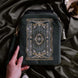 On a sage green background surrounded by flowers sits the Sage green Literati eReader sleeve. it features a green, cream and gold floral embroidered design, a wrist strap and a sage green tassel