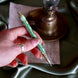 Detail image of the sage green Bibliophile pen by The Quirky Cup Collective. The light green pen features a classic book inspired design in shades of green, white and cream with gold detailing. The pen is held in a hand over a soft green fabric background.