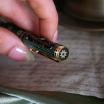 Detail image of the cap of the sage green Bibliophile pen by The Quirky Cup Collective. The light green pen features a classic book inspired design in shades of green, white and cream with gold detailing. The top of the pen cap features a small flower motif.