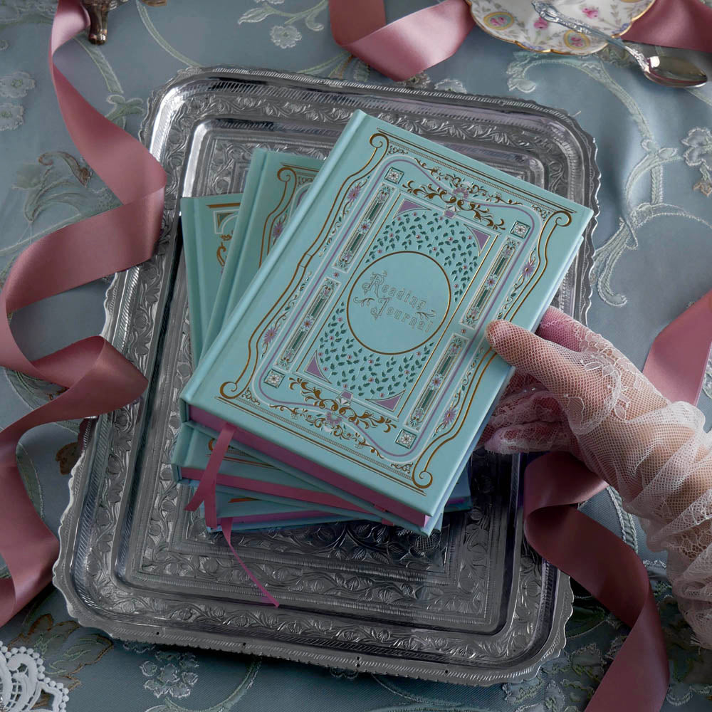 A stack of aqua Antionette journals and reading journals sit on a silver tray on a light blue tablecloth. it's intricate foil covers in shades of pink, gold and blue reference the designs of classic book aesthetic for the perfect whimsical romance book journal.
