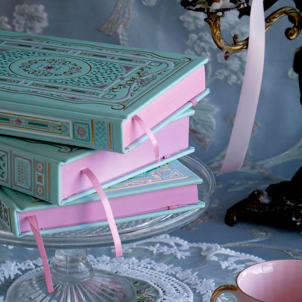 The 3 mint green Antionette journals and reading journals sit together on a class cake stand surrounded by candles, pearls and pink features. The intricate foil covers in shades of pink, gold and blue reference the designs of classical romance books aesthetic with the pink painted edges further providing a luxury book journaling aesthetic.