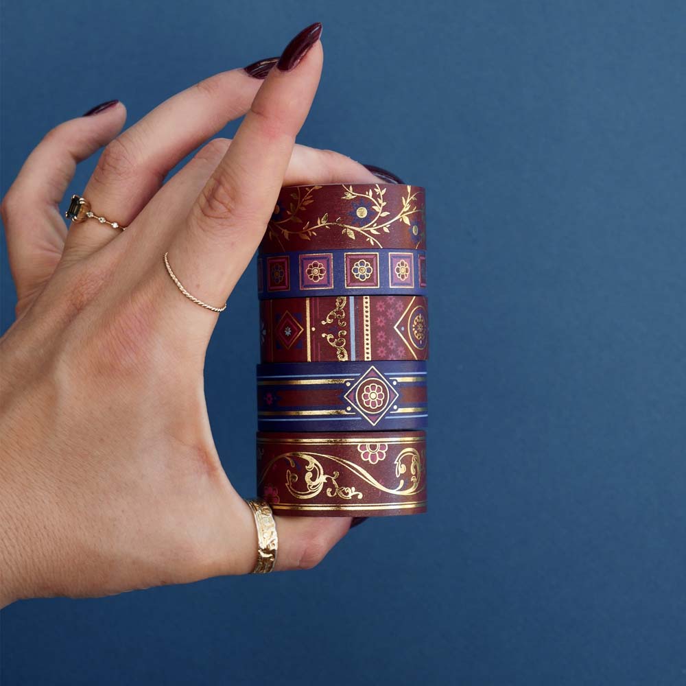 The Crimson variations of the literati  washi tape. The romantic washi tape design is held in a stack by a hand in front of a blue background and is in tones of deep red, royal blue and foiled gold. The patterns vary from geometric to floral and all have a vaguely classical aesthetic.