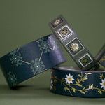 A close up of full set of the Sage green variations of the Literati washi tape by The Quirky Cup Collective sit as rolls in piled stack in a green background. The classic book inspired washi tape designs feature classical book inspired designs and ornamentation in shades of dark green, sage green, white and foiled gold.