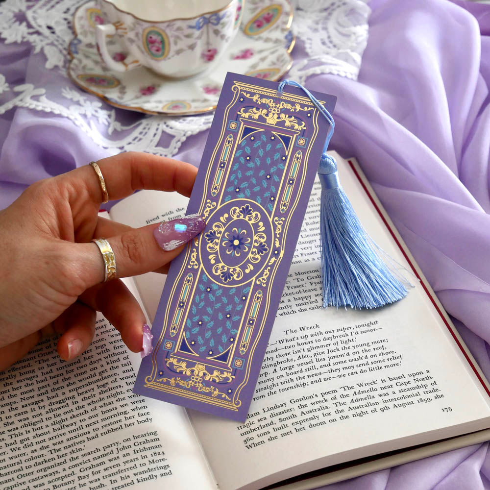 Decorative purple bookmark with tassel held over an open book on a lilac fabric background.