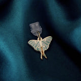 A Luna Moth dust plug design by The Quirky Cup Collective sits on a deep green fabric background. The gold Night luna moth design (as part of the romantasy kindle decor release) featured green detailing.