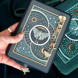 A detail of the Luna Moth eReader decor bundle. On a deep green background a hand holds a kindle with the Luna moth  eReader sticker insert and dust plug. It sits on the matching eReader case and has a matching charmlet attached. The range is all in a shade of teal and features gold, cream and mint detailing and embroidery.