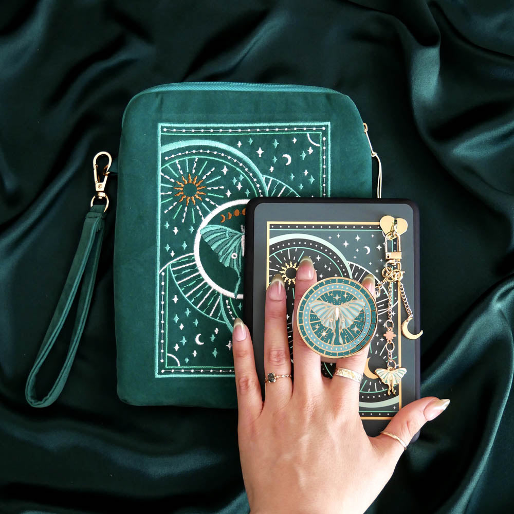 An image of the Luna Moth eReader decor bundle on a deep emerald fabric background. The set features an eReader sleeve, eReader sticker insert, charmlet, clingie (like a pop socket!) and dust plug all in the same teal colour with mint, gold and white detailing. The intricate Luna moth design features star and moon motifs. The eReader case features a wrist strap and gold tassel.
