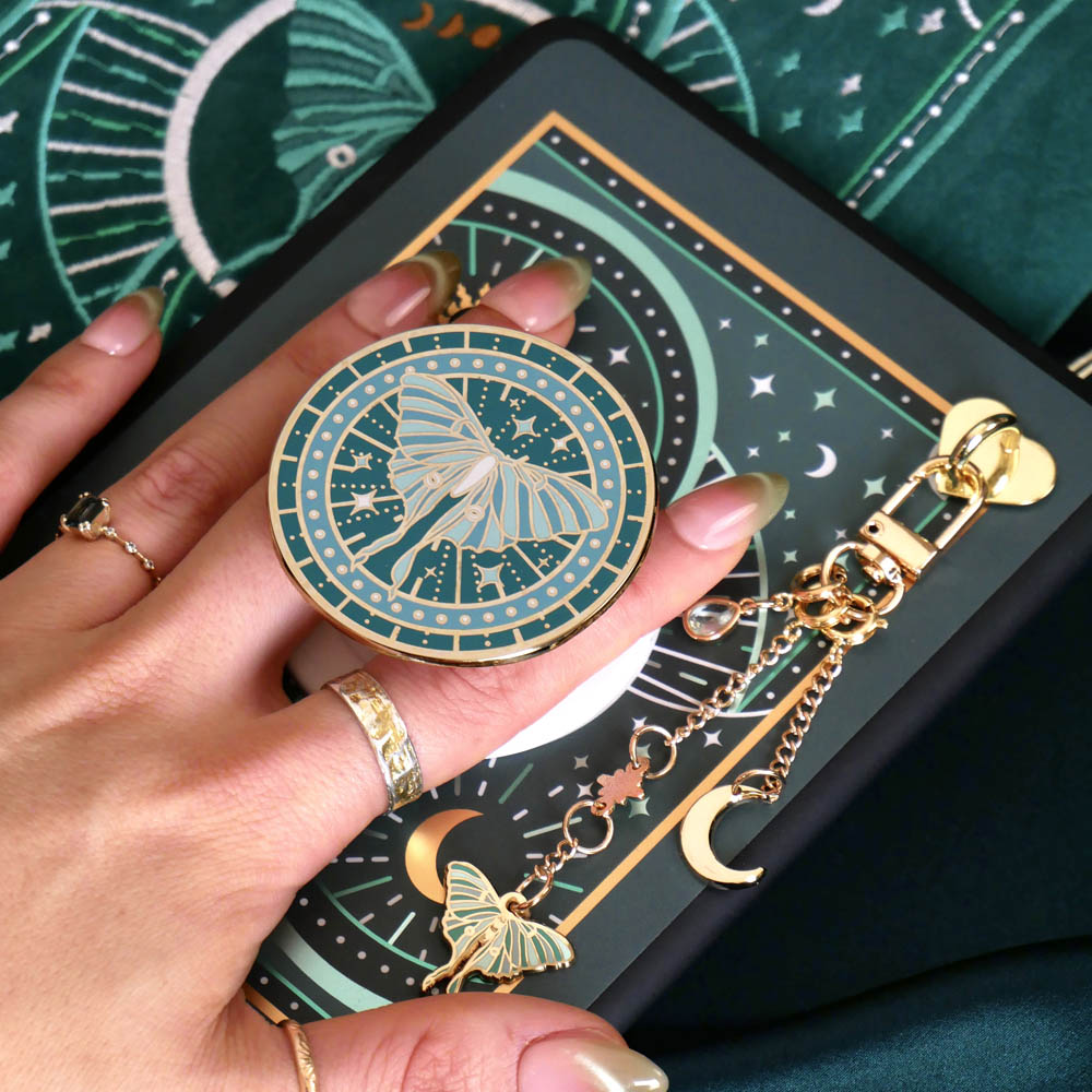 A detail of the Luna Moth eReader decor bundle. On a deep green background a hand holds the Luna moth clingie (think an enamel pop socket!), attached to a kindle with matching eReader sticker insert. It sits on the matching eReader case and has a matching charmlet attached. the range is all in a shade of teal and features gold, cream and mint detailing and embroidery.