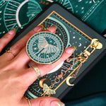 A detail of the Luna Moth eReader decor bundle. On a deep green background a hand holds the Luna moth clingie (think an enamel pop socket!), attached to a kindle with matching eReader sticker insert. It sits on the matching eReader case and has a matching charmlet attached. the range is all in a shade of teal and features gold, cream and mint detailing and embroidery.
