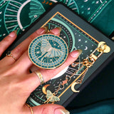 A detail of the Luna Moth eReader decor bundle. On a deep green background a hand holds the Luna moth clingie (think an enamel pop socket!), attached to a kindle with matching eReader sticker insert. It sits on the matching eReader case and has a matching charmlet attached. the range is all in a shade of teal and features gold, cream and mint detailing and embroidery.