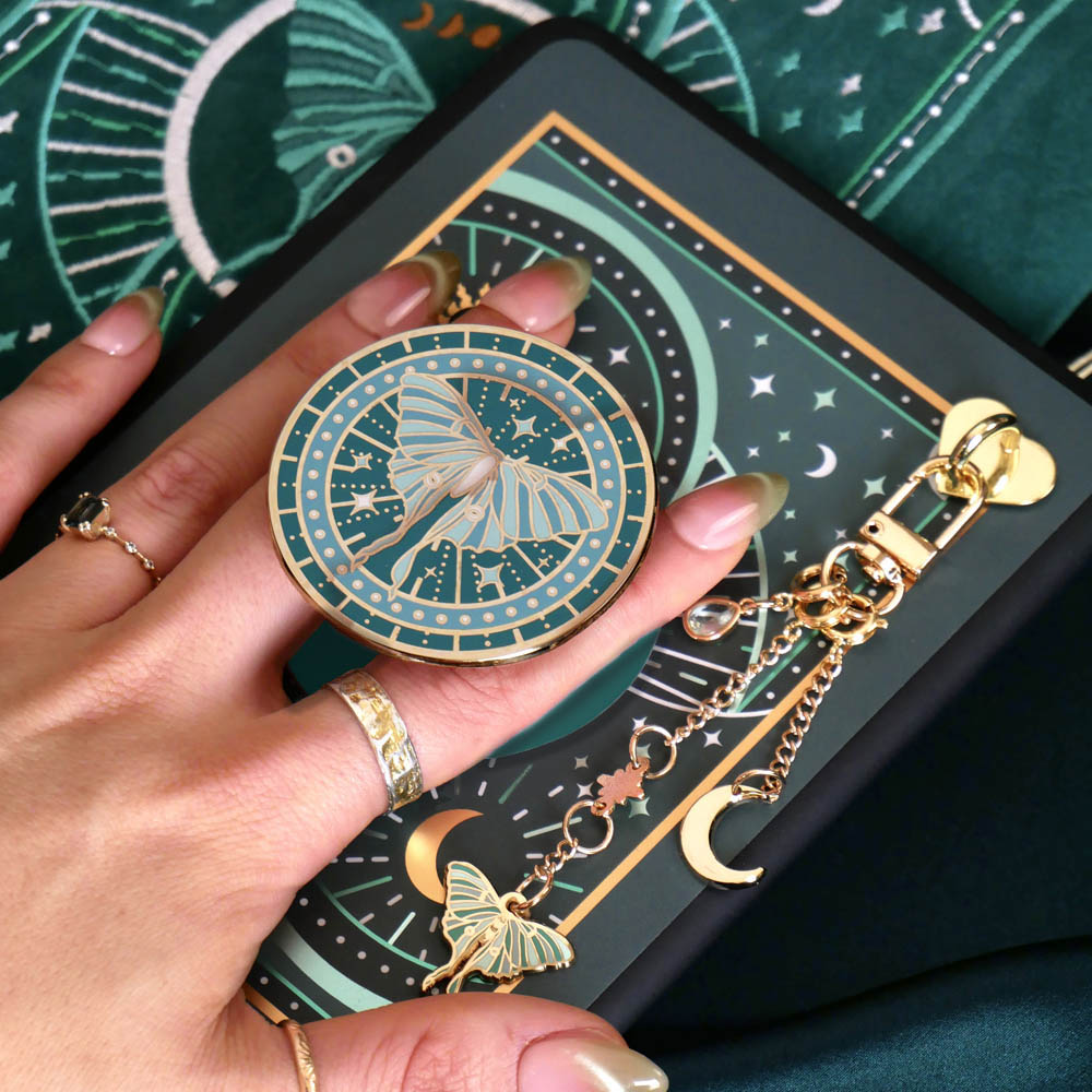 A close-up of a hand holding a round celestial-themed magnetic kindle grip with a luna moth charm attached by a chain. The design features teal, gold, and cream moon and star motifs, with a matching patterned surface visible in the background. By The Quirky Cup Collective